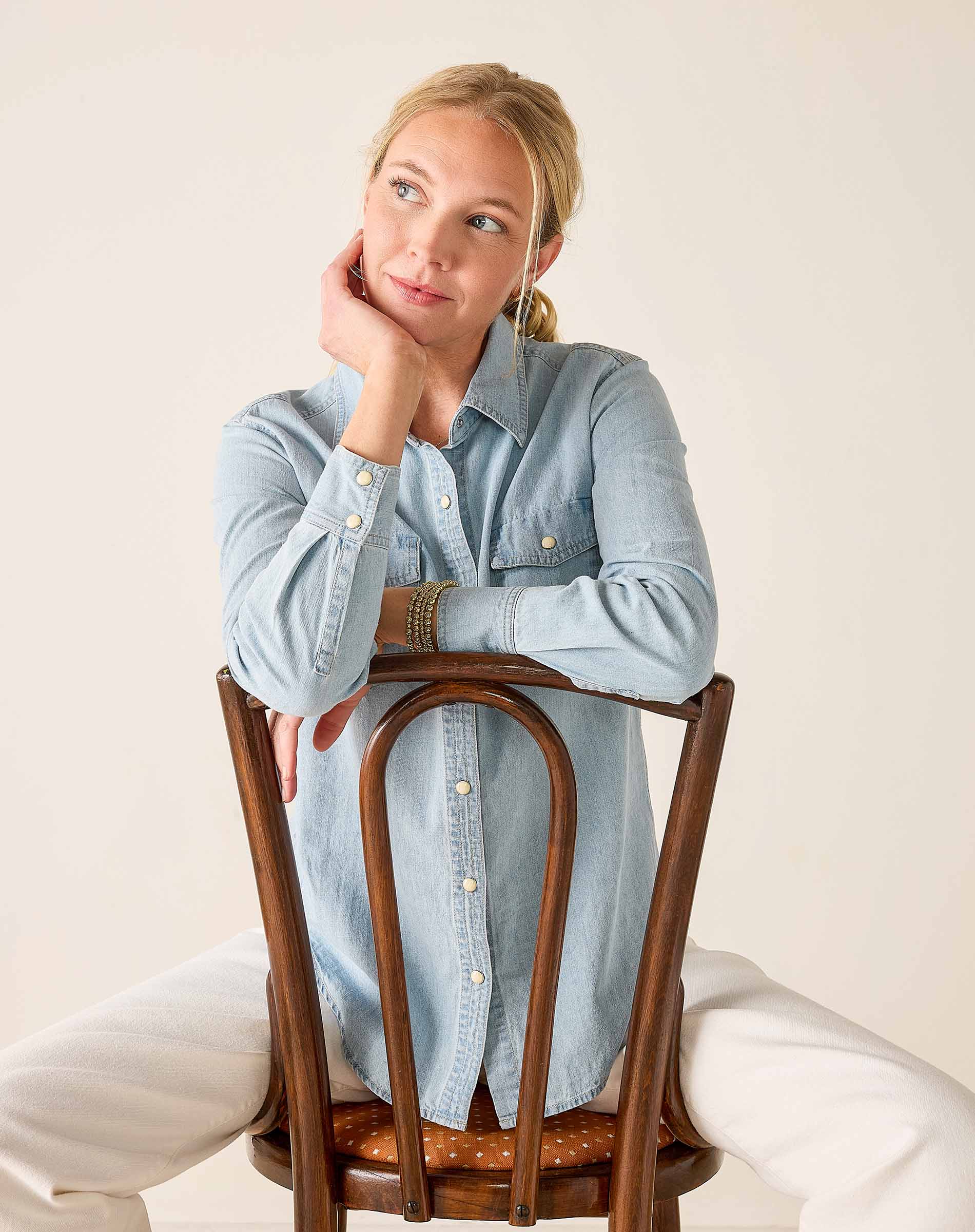 Woman sitting on a wooden chair wearing a light blue denim shirt against a plain background