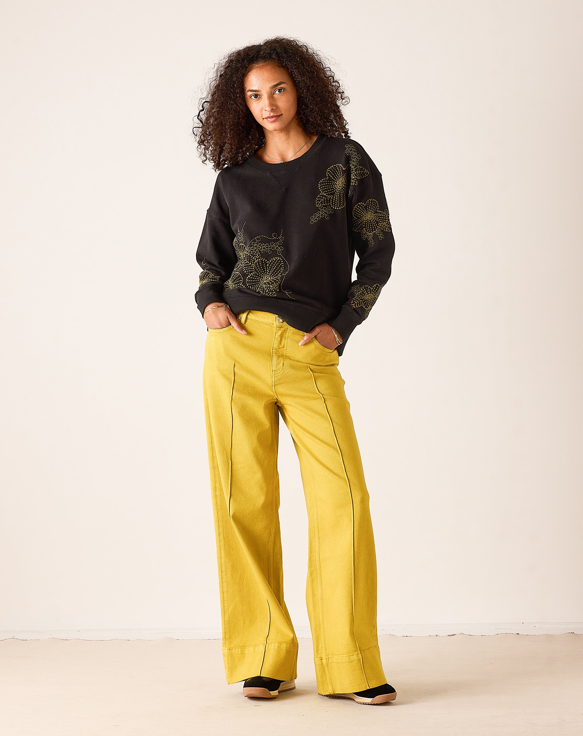 Dark charcoal crewneck sweatshirt with large yellow and white floral embroidery, paired with mustard yellow wide-leg trousers.