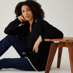 woman showcasing mersea marina polo sweater in black with sand color detail sitting with arm on stool