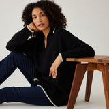 woman showcasing mersea marina polo sweater in black with sand color detail sitting with arm on stool