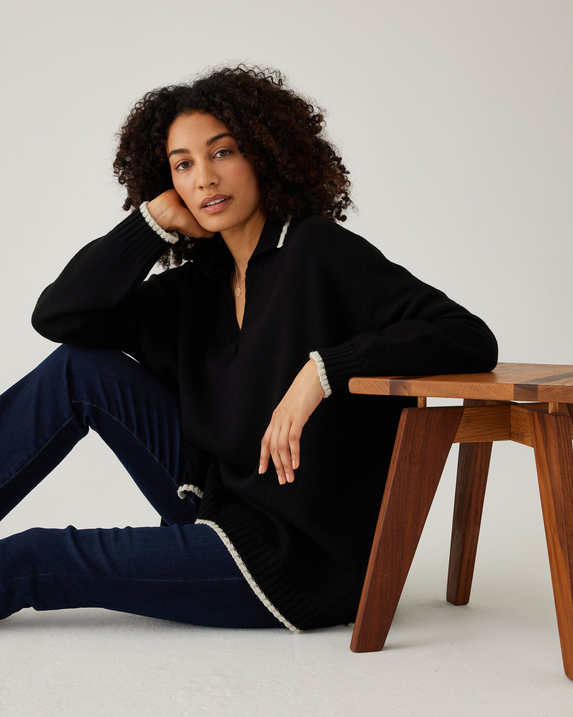 woman showcasing mersea marina polo sweater in black with sand color detail sitting with arm on stool