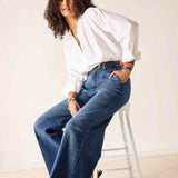 Woman wearing a white blouse and blue jeans sitting on a stool against a plain background