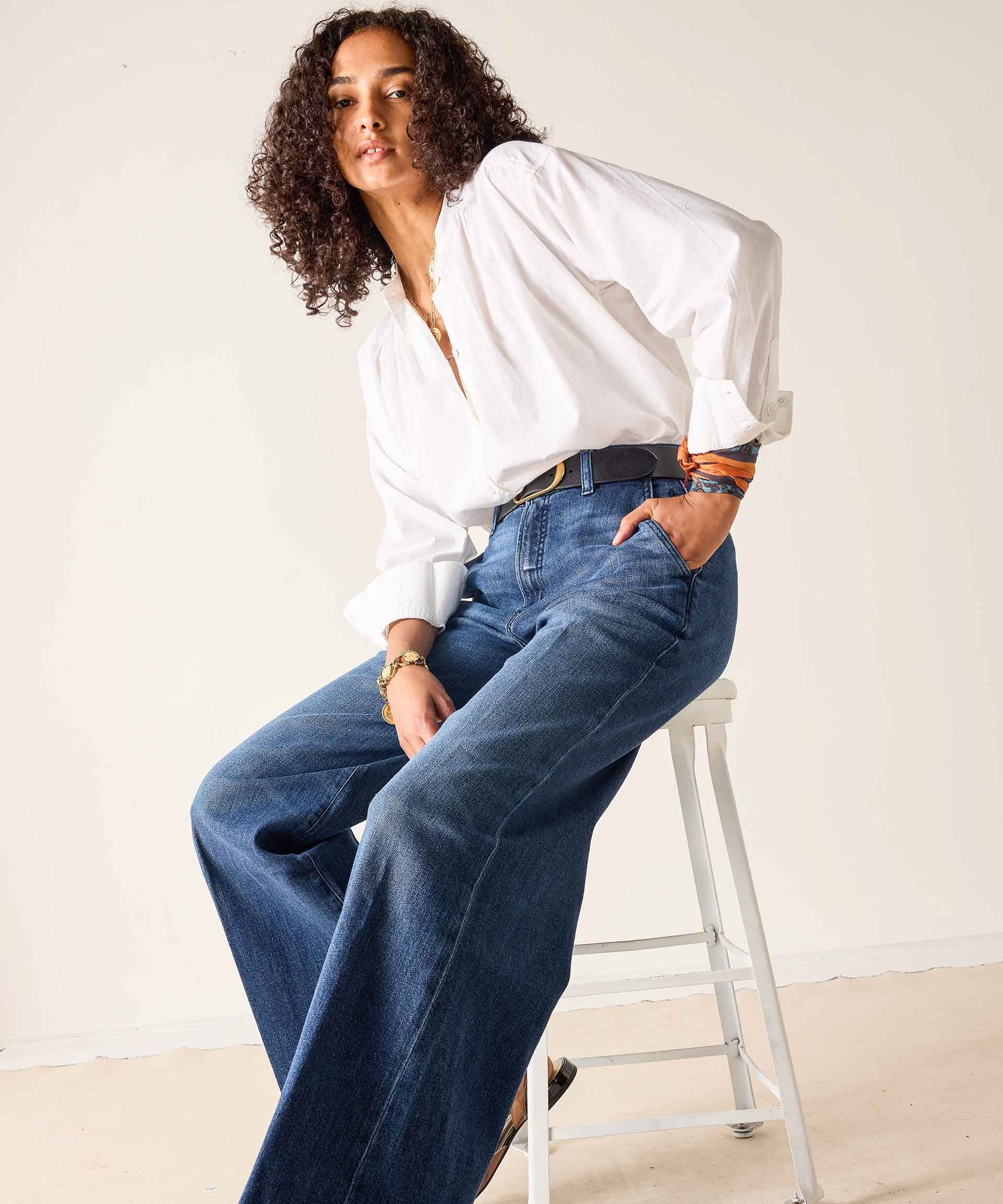 Woman wearing a white blouse and blue jeans sitting on a stool against a plain background
