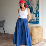 Woman wearing a white tank top and blue wide-leg pants with a red headscarf, standing in a room with a textured wall and wicker chair.
