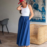 Woman wearing a white top and blue skirt with a red headscarf, standing in a room with a wicker chair and abstract painting.