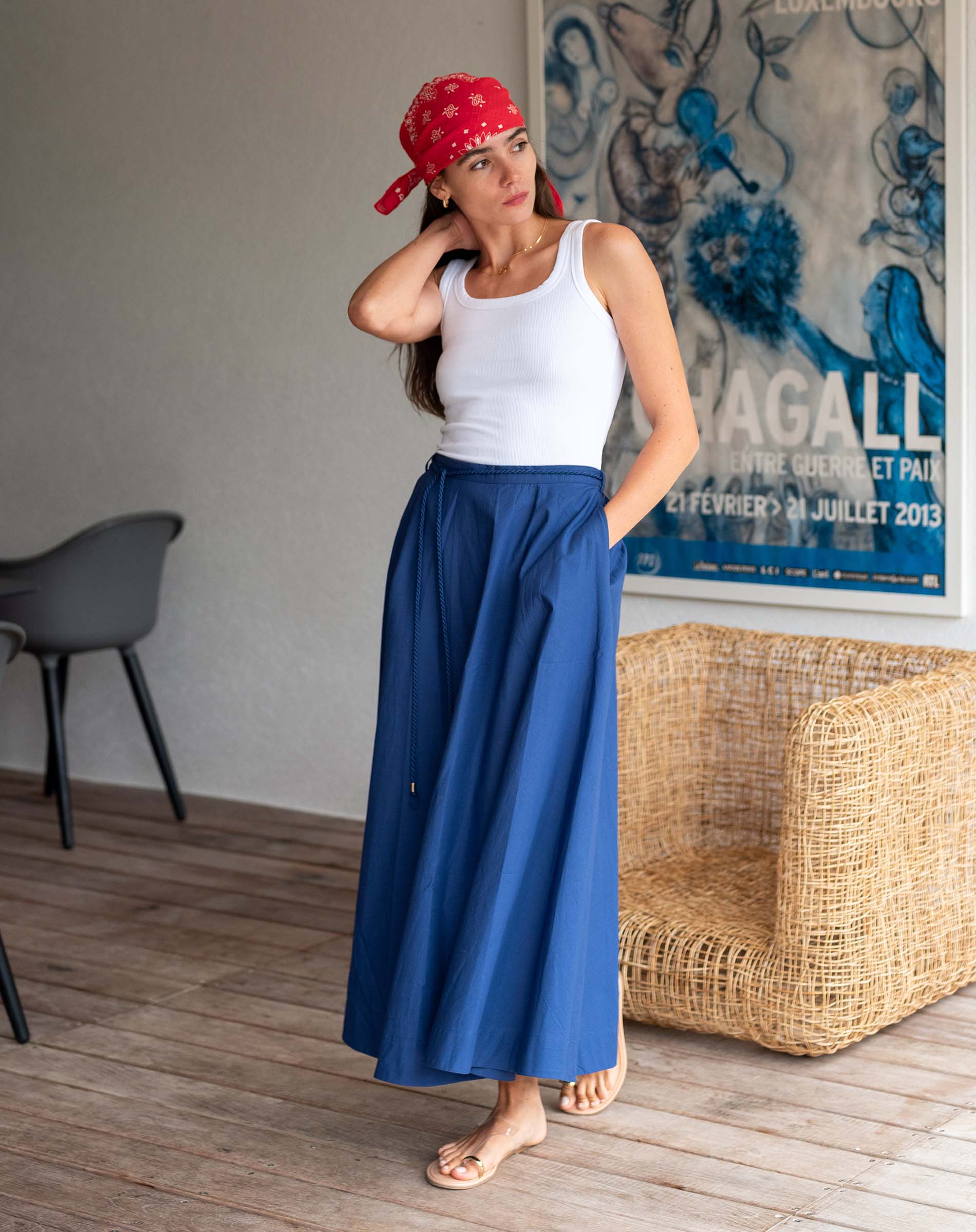 Woman wearing a white top and blue skirt with a red headscarf, standing in a room with a wicker chair and abstract painting.