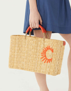female holding large straw basket with leather orange handles and an orange sun on white background