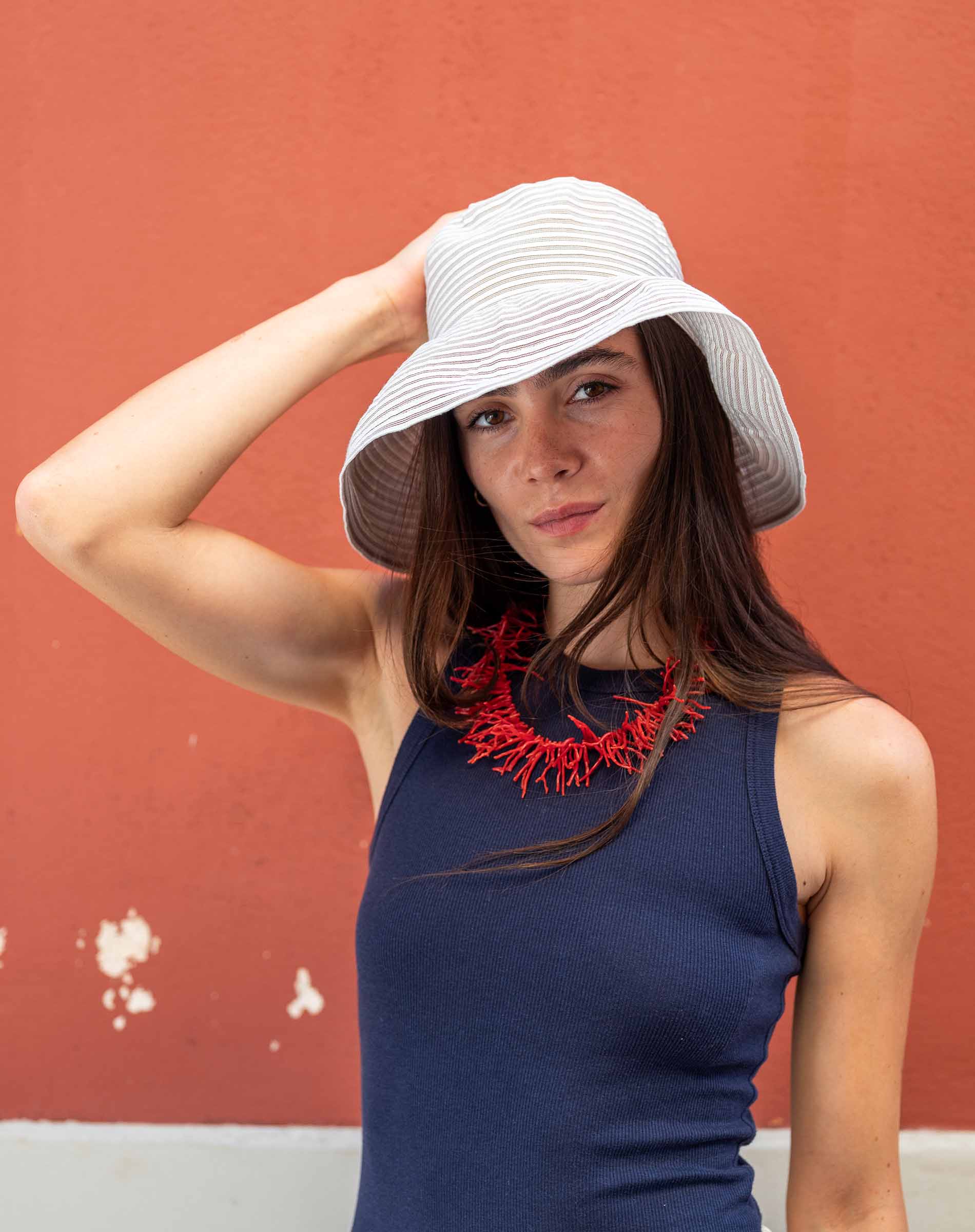 Woman wearing a white sun hat and blue dress with a red necklace against a red wall.