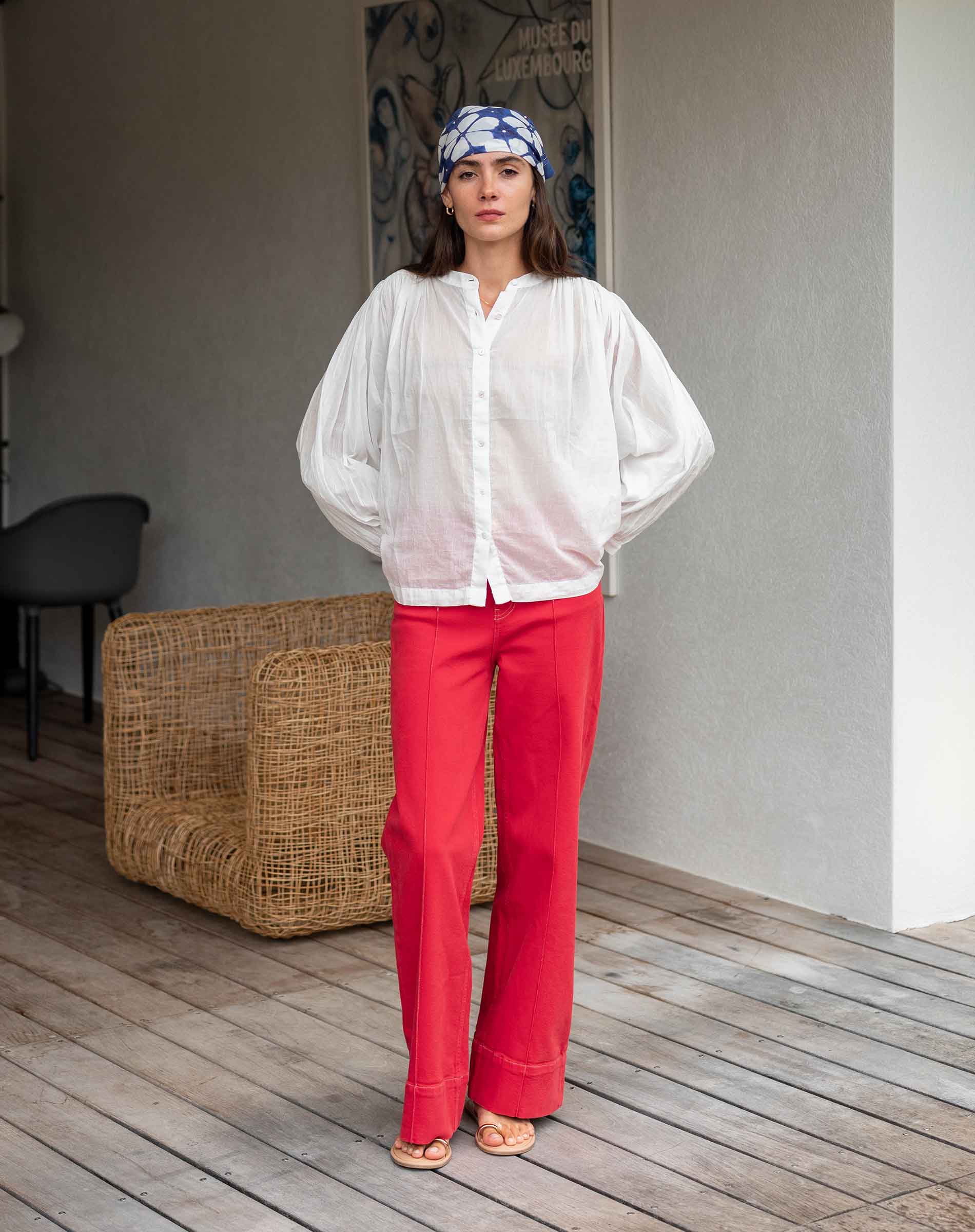 Woman wearing a white blouse and red pants standing in a room with a textured wall and wicker basket.