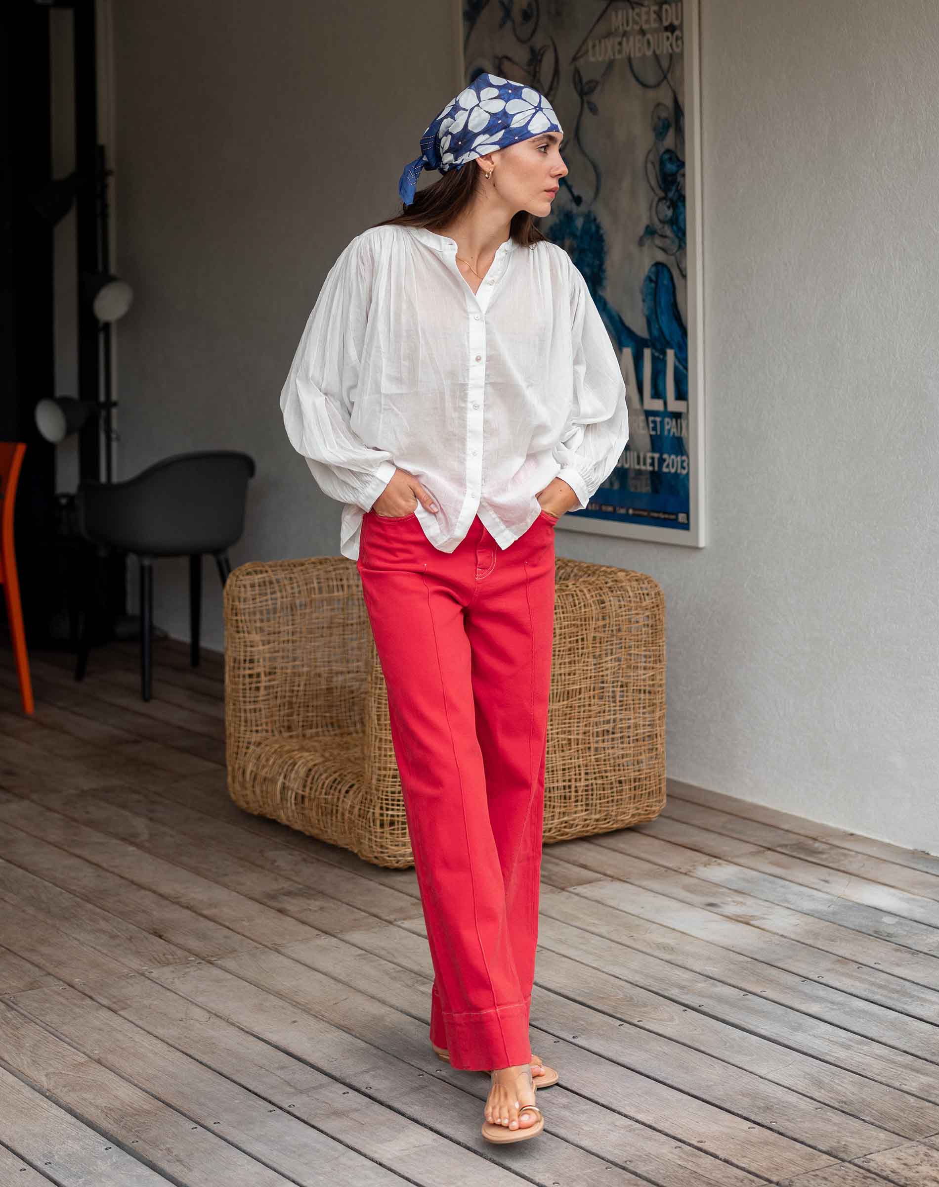 Woman wearing a white blouse and red pants with a blue patterned headscarf indoors.