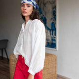 Woman wearing a white blouse and red pants with a blue headscarf, standing in a room with a wicker chair and a poster on the wall.