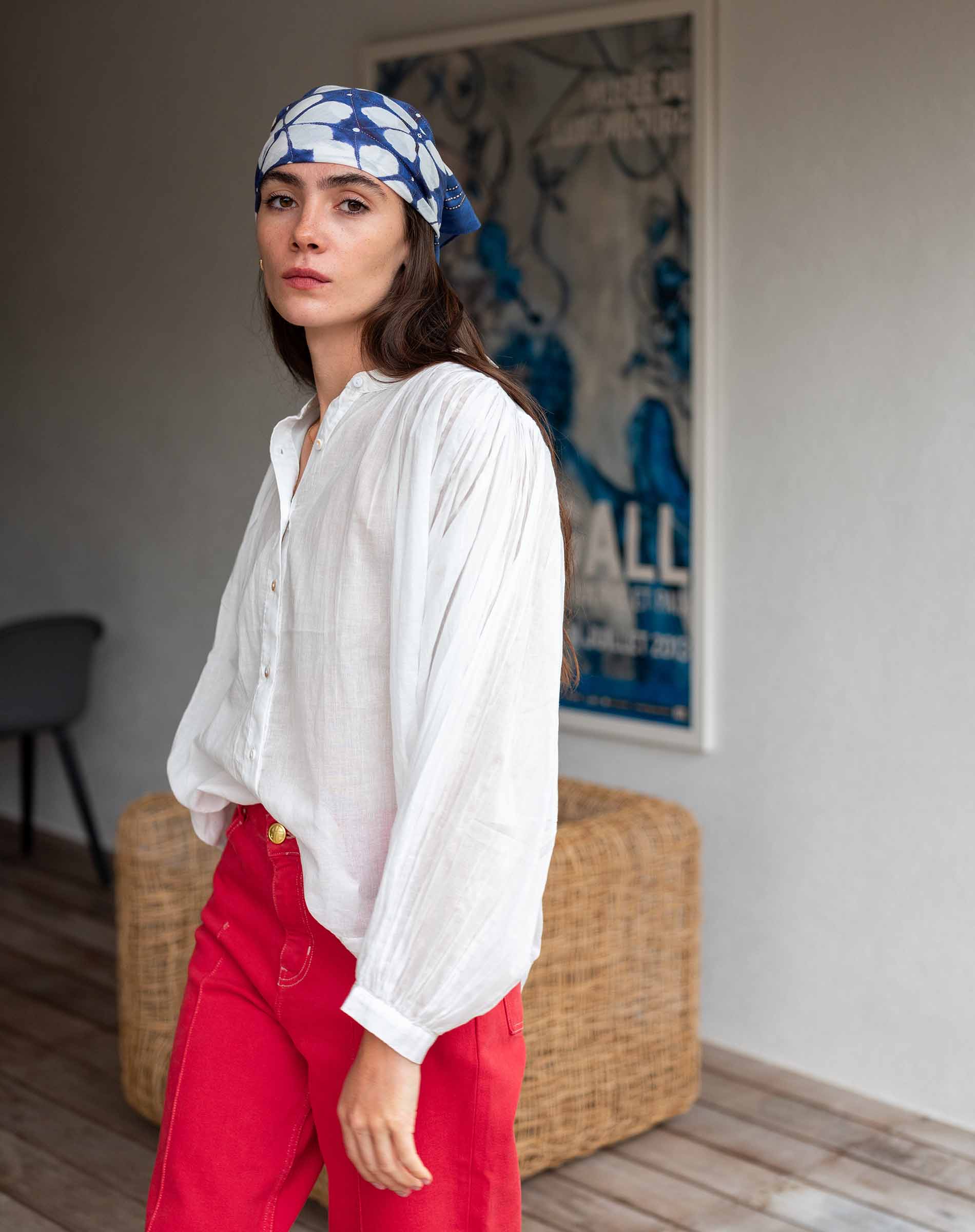 Woman wearing a white blouse and red pants with a blue headscarf, standing in a room with a wicker chair and a poster on the wall.
