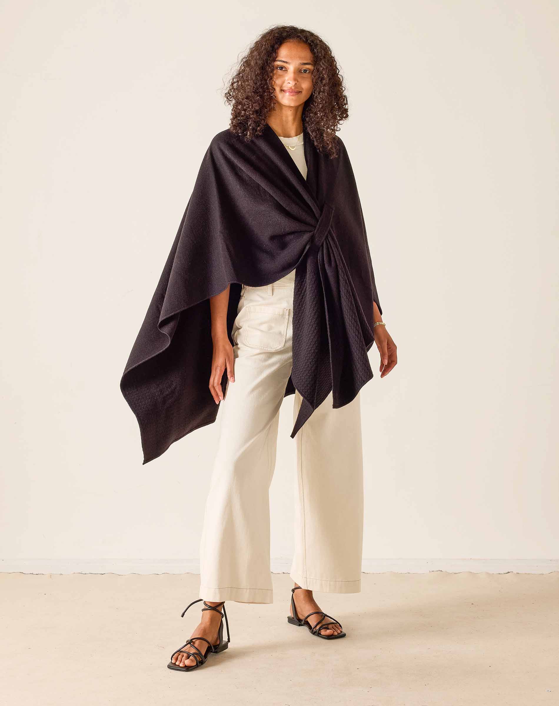 Woman wearing a black wrap over a white top and pants on a plain background