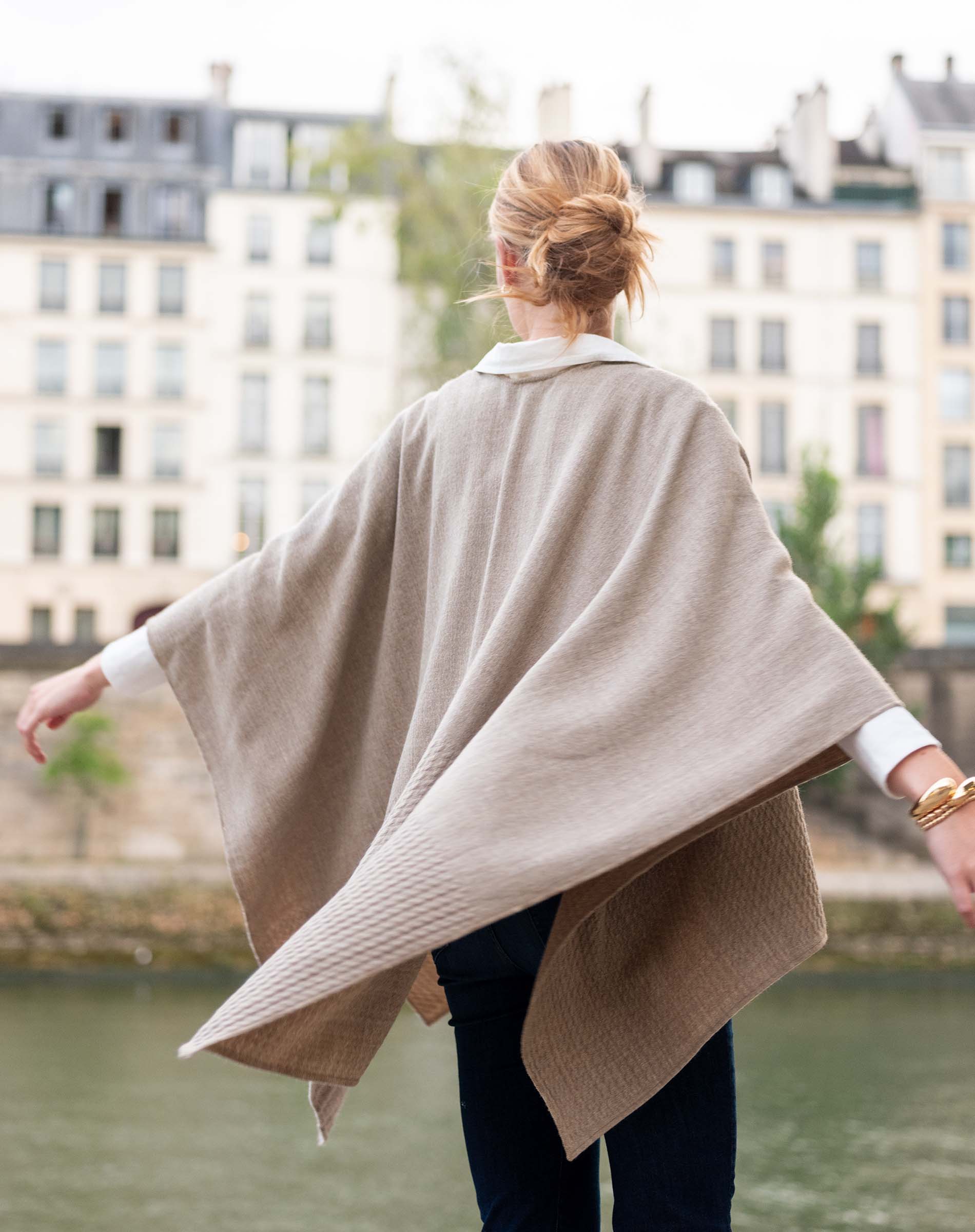 Person wearing a beige poncho in an urban setting with buildings and water in the background