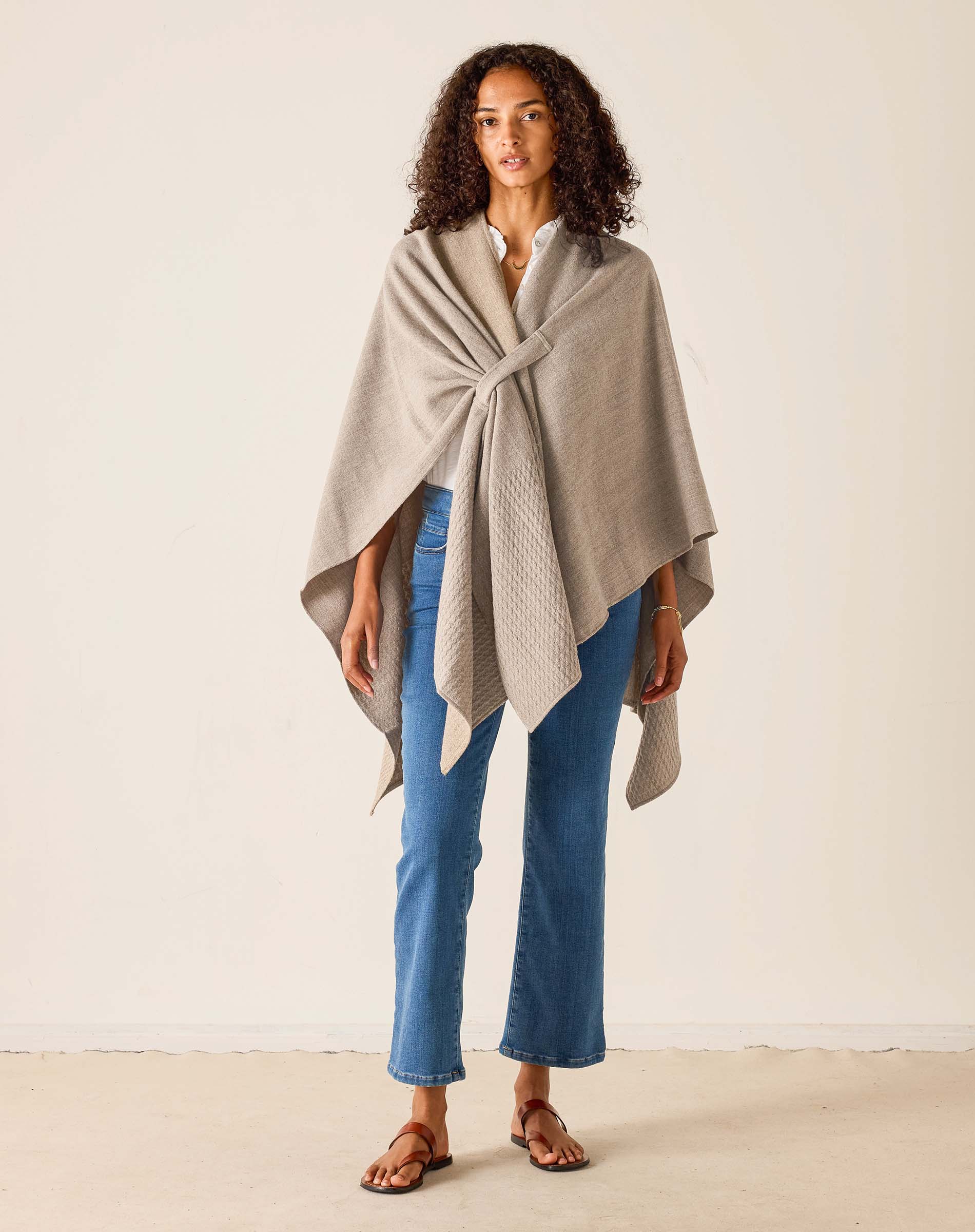 Woman wearing a beige poncho over a white shirt with blue jeans on a plain background