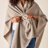 Woman wearing a beige poncho over a white shirt and blue jeans on a plain background