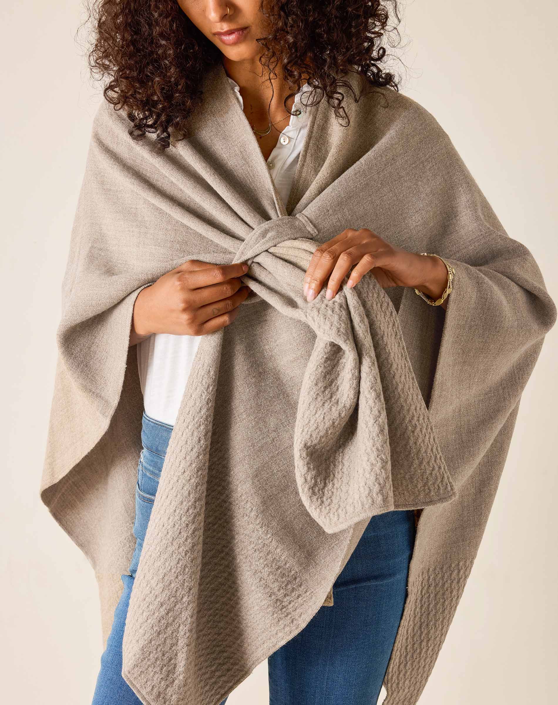 Woman wearing a beige poncho over a white shirt and blue jeans on a plain background