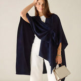Woman wearing a navy blue poncho over a white outfit, holding a beige handbag against a plain background.