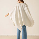 Woman wearing a white poncho and blue jeans against a beige background
