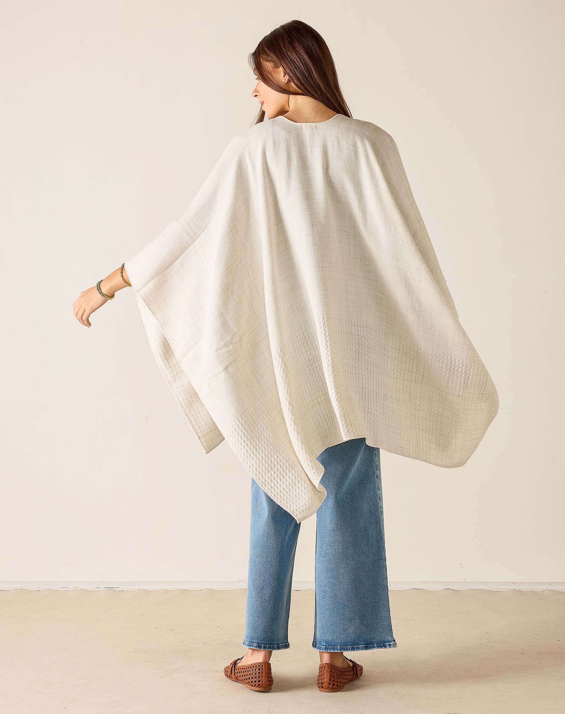 Woman wearing a white poncho and blue jeans against a beige background