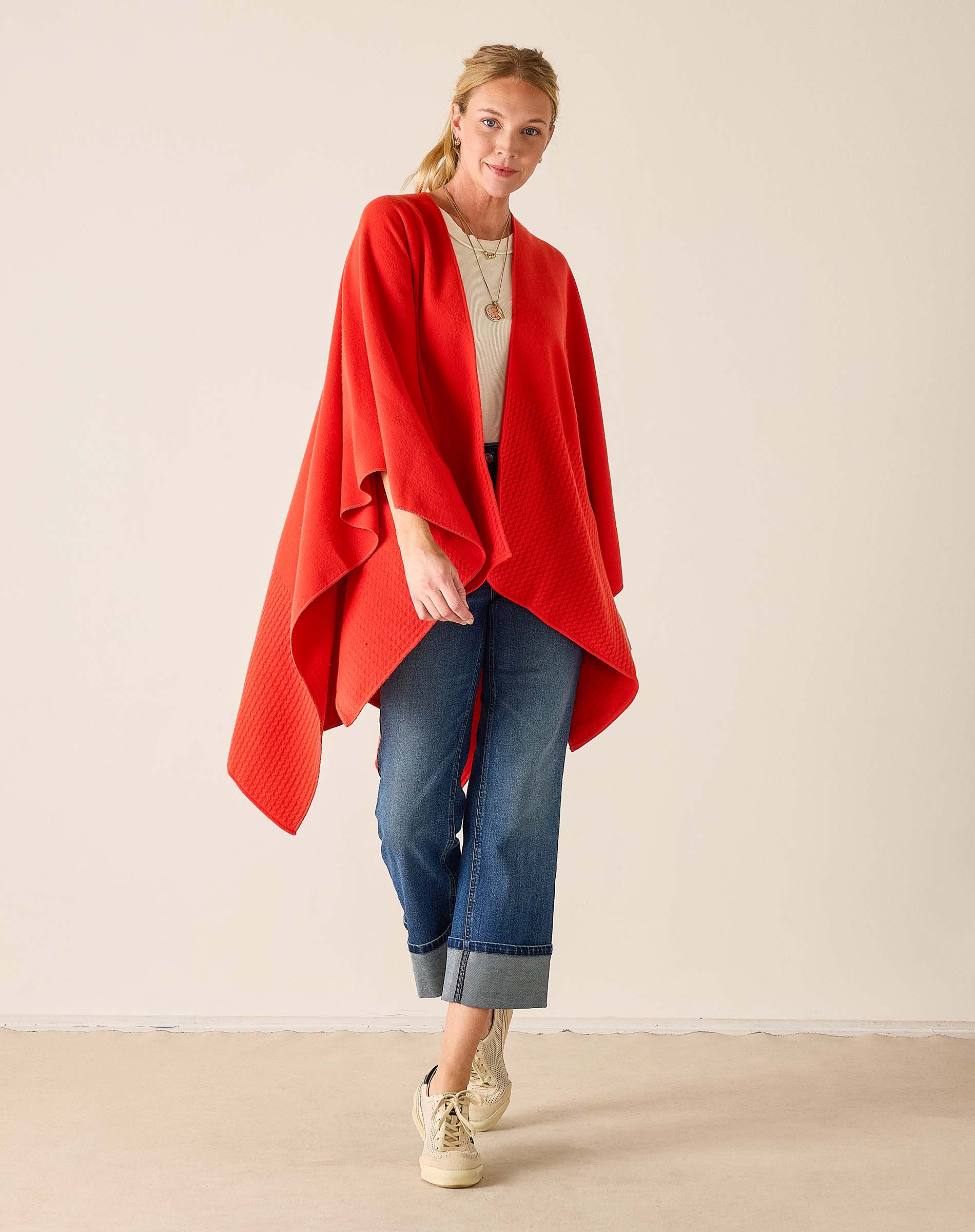 Woman wearing a red poncho, white shirt, blue jeans, and beige sneakers on a plain background