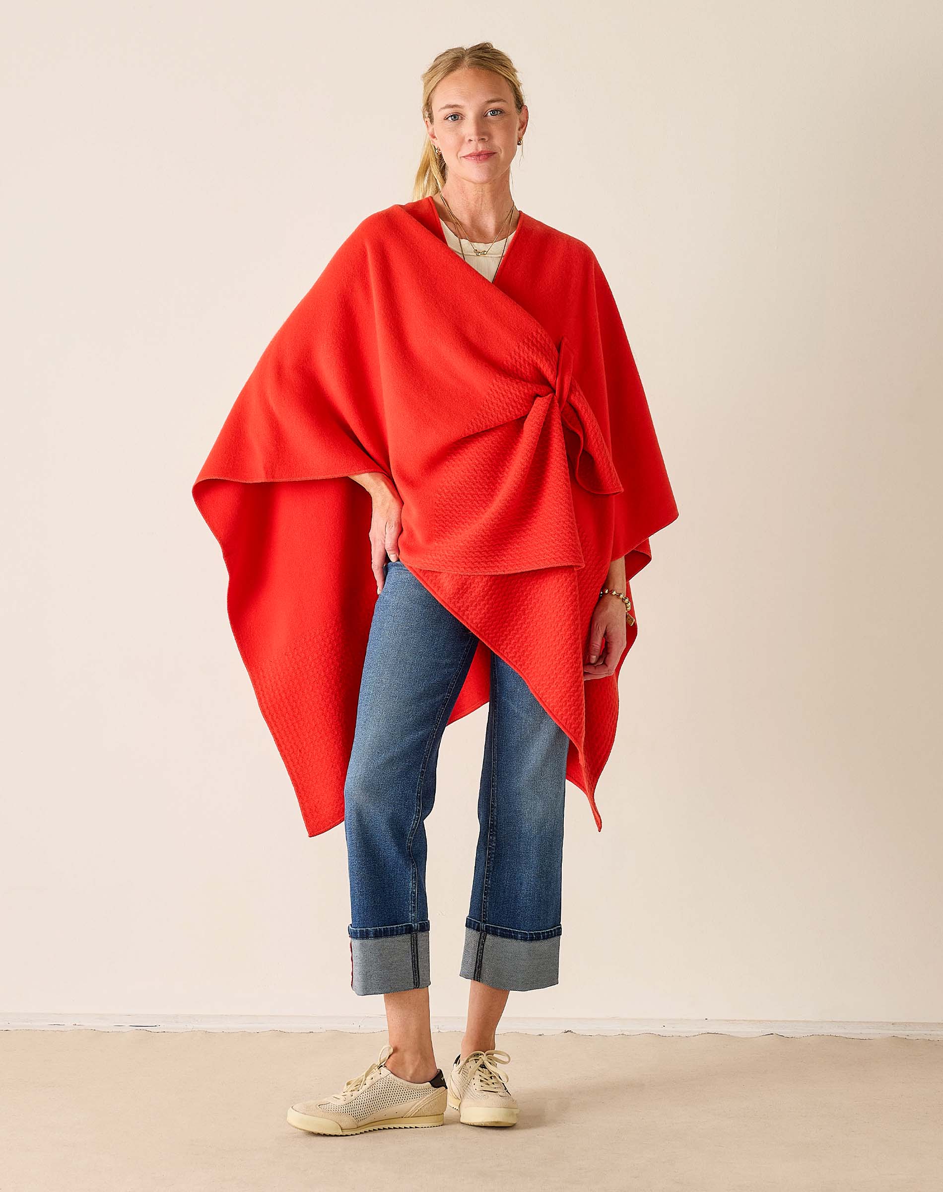Woman wearing a red poncho over a white shirt and blue jeans on a beige background