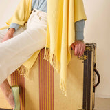 Person sitting on a vintage suitcase with a yellow blanket draped over them, wearing a blue shirt and white pants.