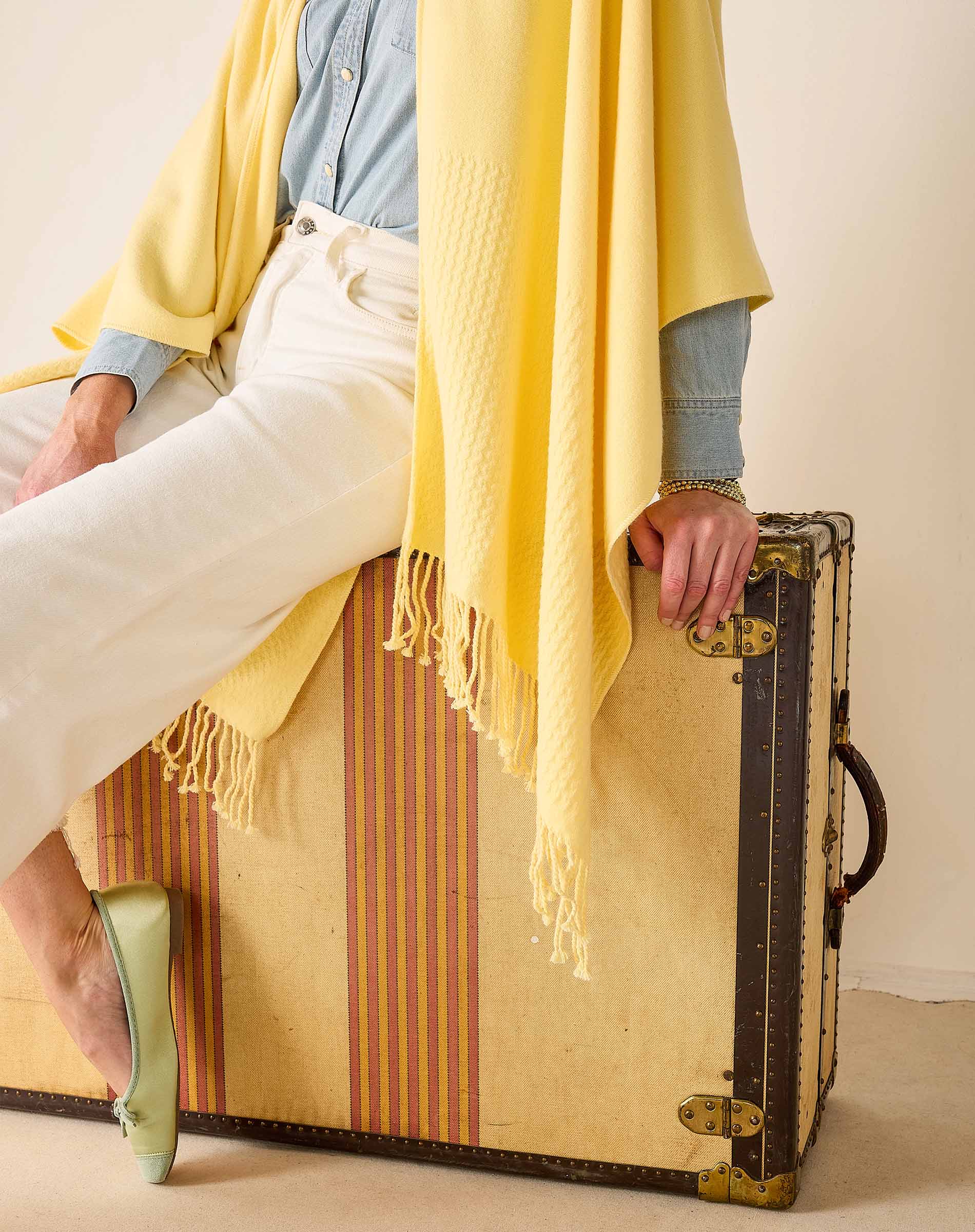 Person sitting on a vintage suitcase with a yellow blanket draped over them, wearing a blue shirt and white pants.