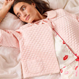 Woman wearing a pink quilted jacket and floral pajama pants lying on a bed.