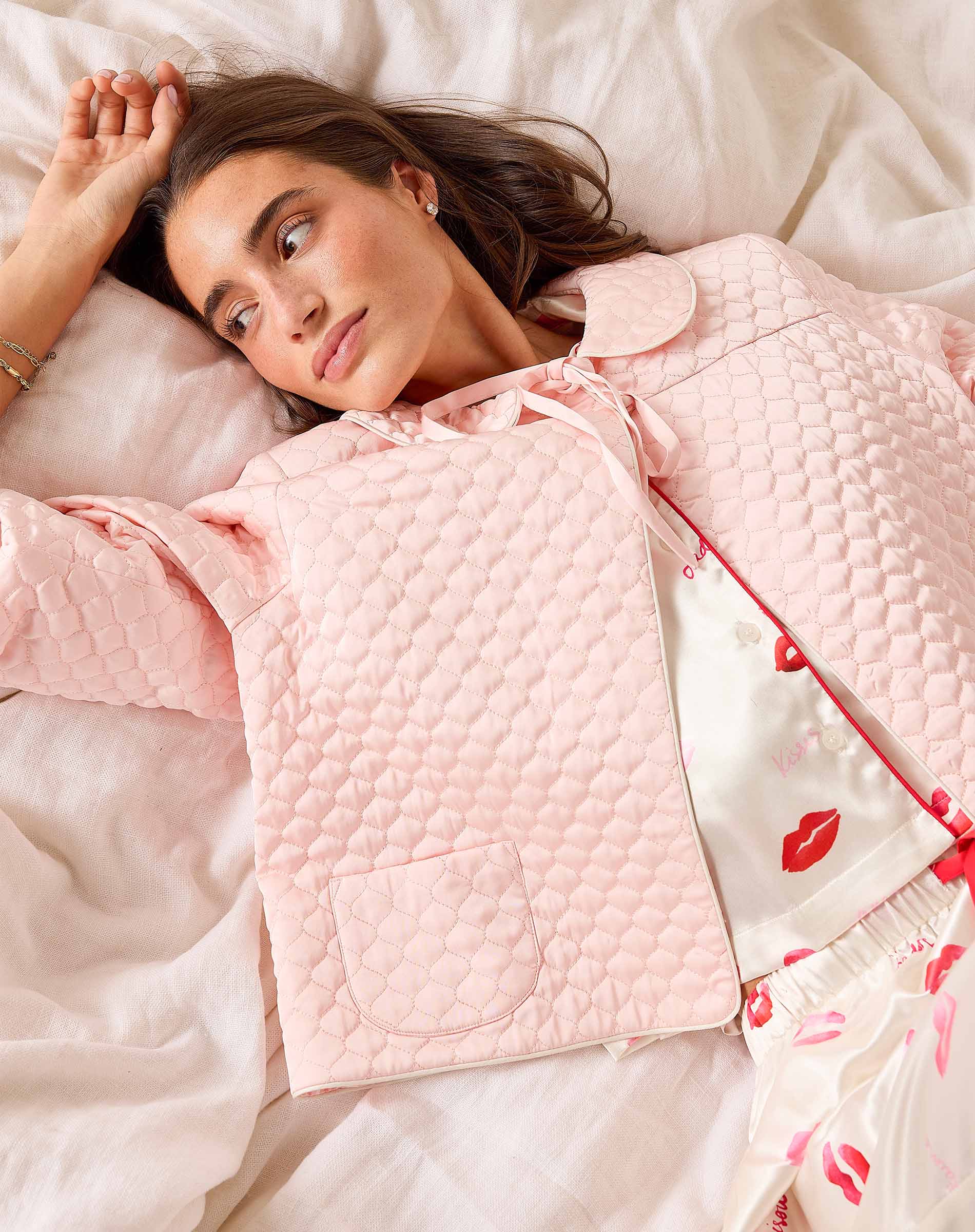 Woman wearing a pink quilted jacket and floral pajama pants lying on a bed.
