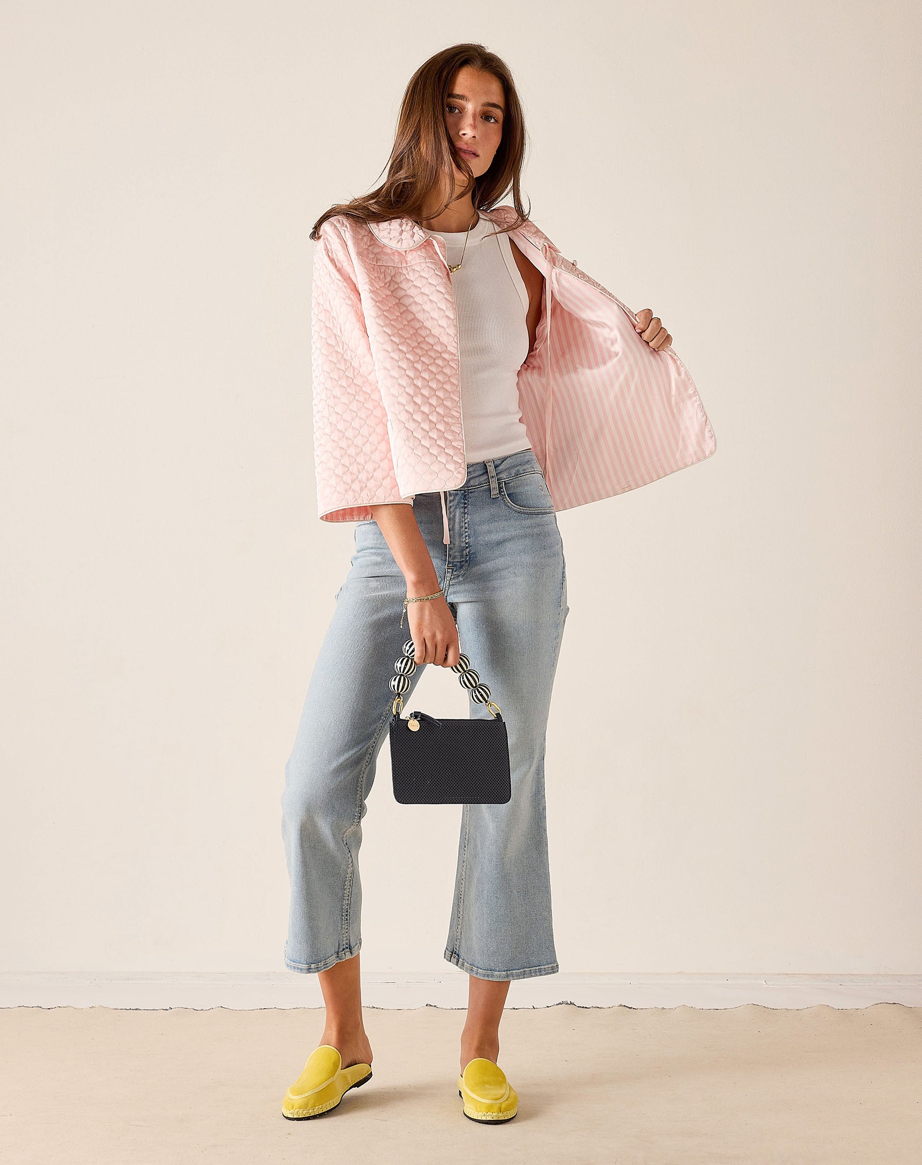 Woman wearing a pink quilted jacket, white shirt, blue jeans, and yellow shoes against a beige background