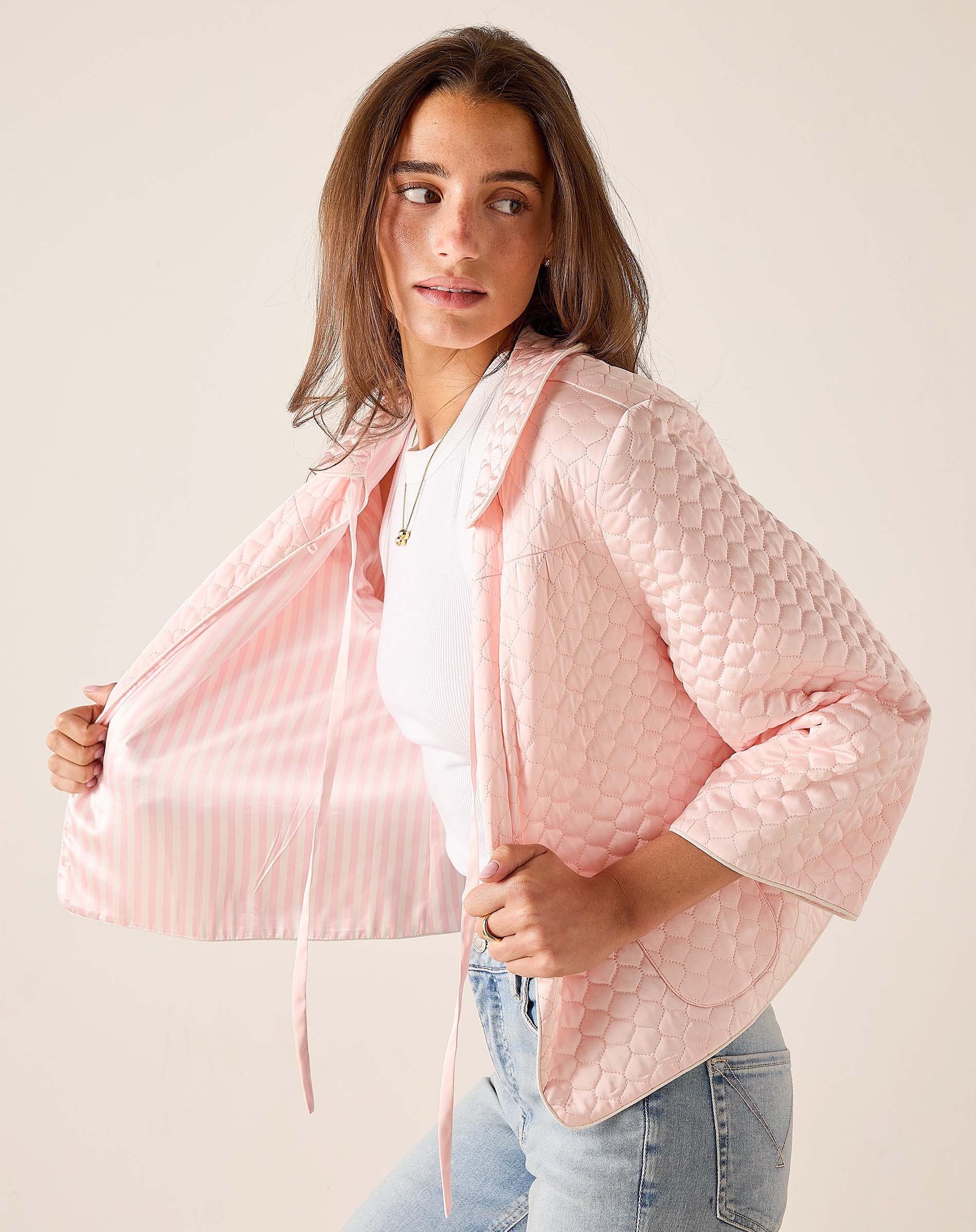 Woman wearing a light pink quilted jacket over a white top and blue jeans on a plain background