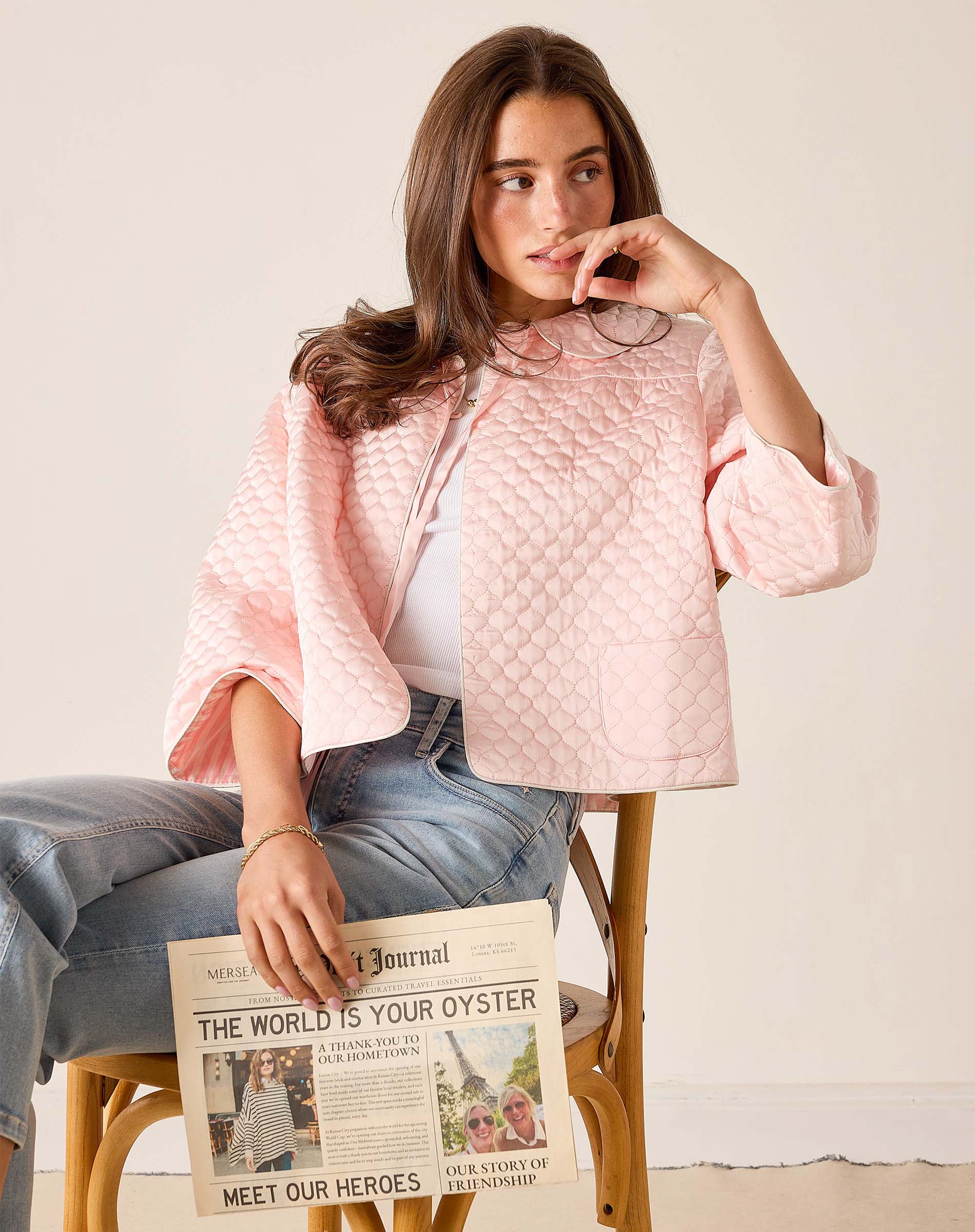 Woman in a pink quilted jacket holding a newspaper on a wooden chair.