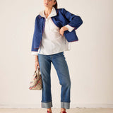 Woman wearing a blue quilted jacket, white shirt, blue jeans, and red shoes on a plain background