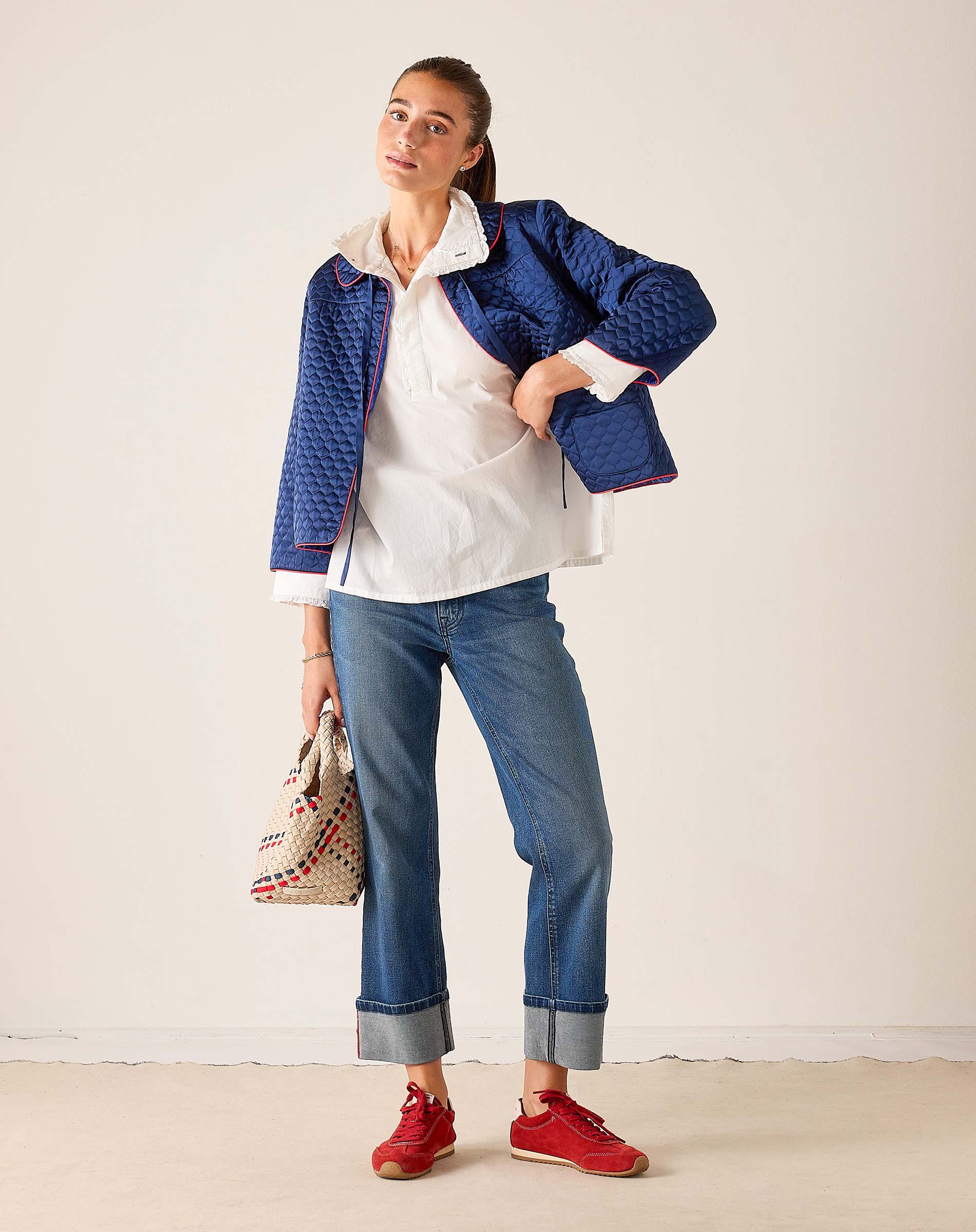 Woman wearing a blue quilted jacket, white shirt, blue jeans, and red shoes on a plain background