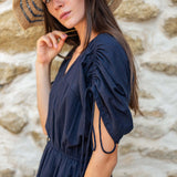 Woman wearing a navy blue dress with cut-out shoulders against a stone wall.