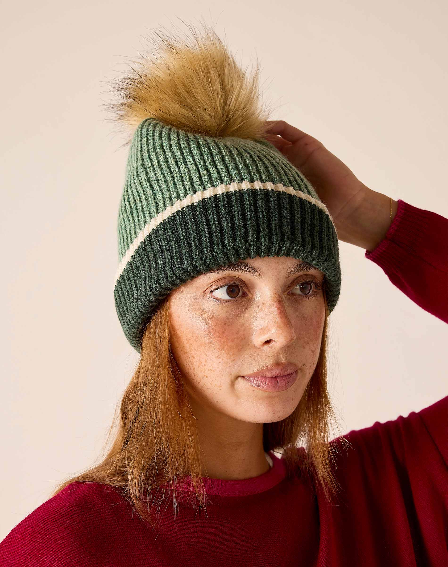 Sage green knit beanie with cream stripe and tan pom-pom.