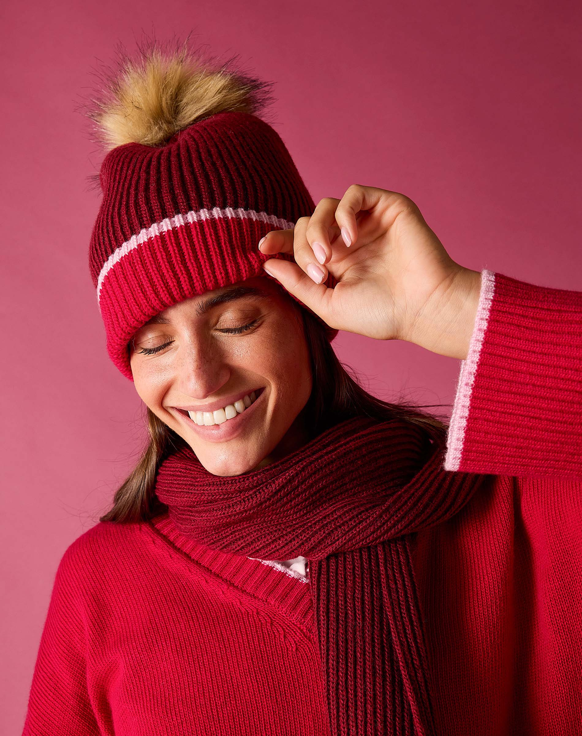 Raspberry pink ribbed knit beanie with a large faux fur pom-pom and white contrast stitching.