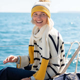 A smiling woman wearing a knit beanie and a cream-colored scarf with yellow accents over a striped sweater sits on a boat against a background of blue ocean water.