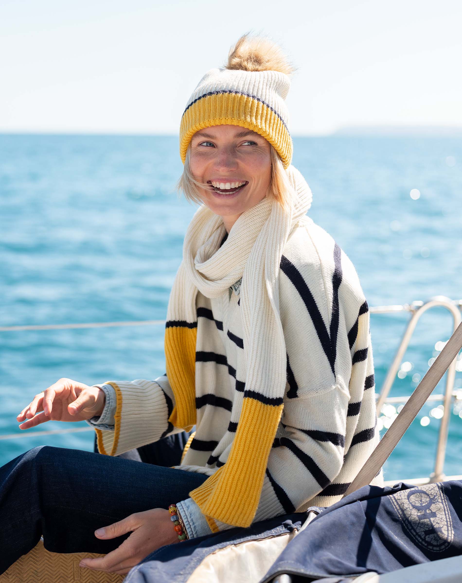 A smiling woman wearing a knit beanie and a cream-colored scarf with yellow accents over a striped sweater sits on a boat against a background of blue ocean water.