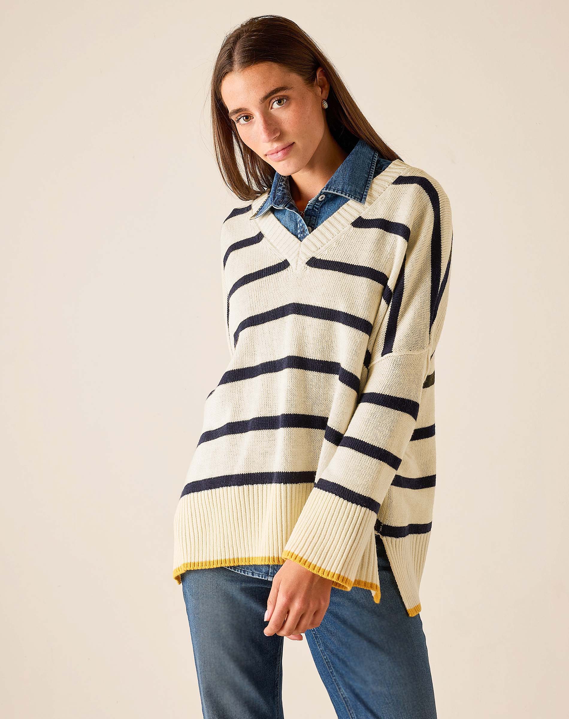 Cream knit v-neck top with navy stripes and a yellow hem over blue jeans.