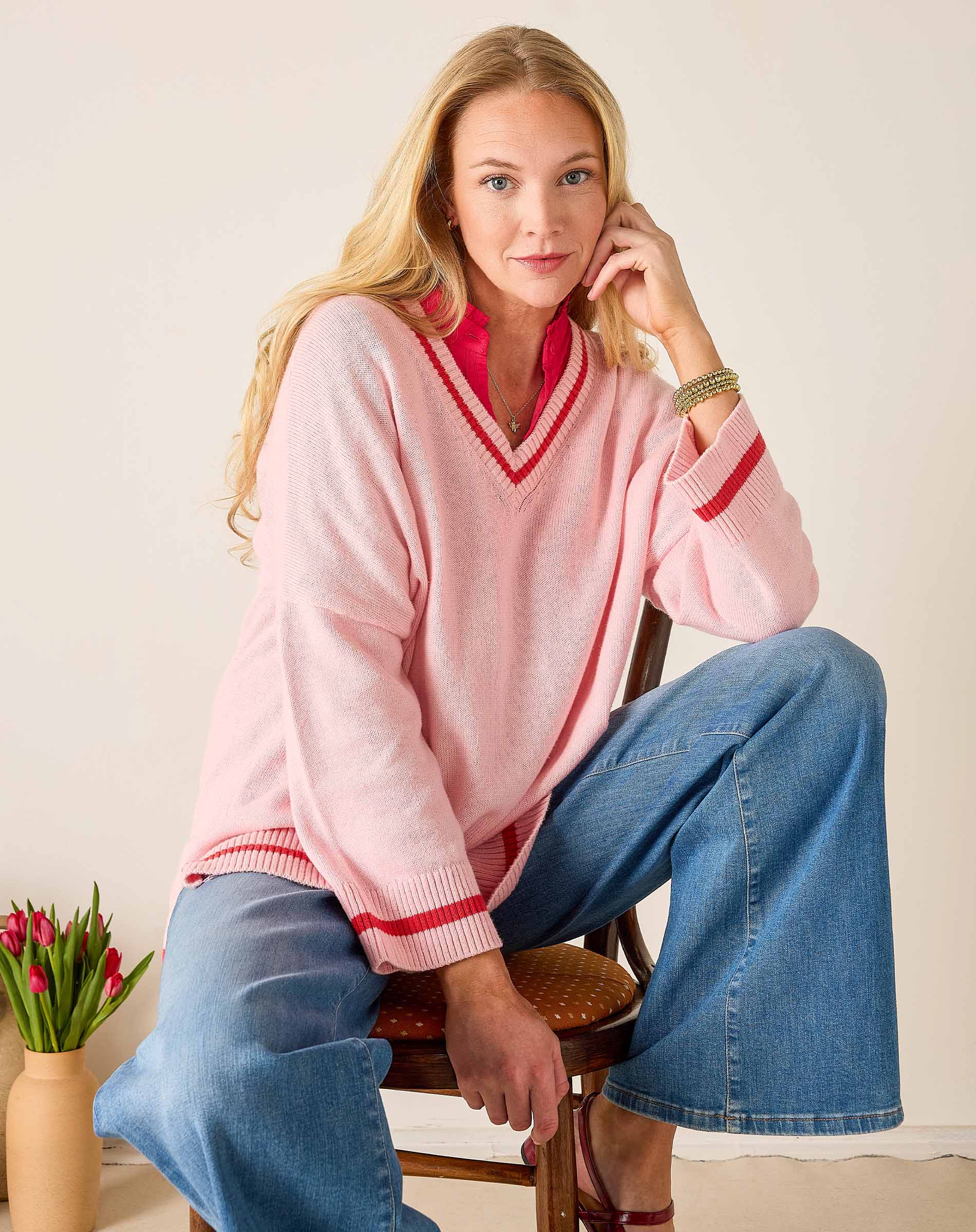 Woman wearing a pink sweater with red trim and blue jeans sitting on a chair.