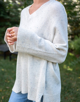 close up of female wearing white speckled v-neck sweater standing outside near trees