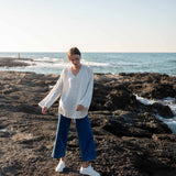 woman wearing mersea montauk v-neck sweater with wide sleeves in cream color standing on rocky beach near water