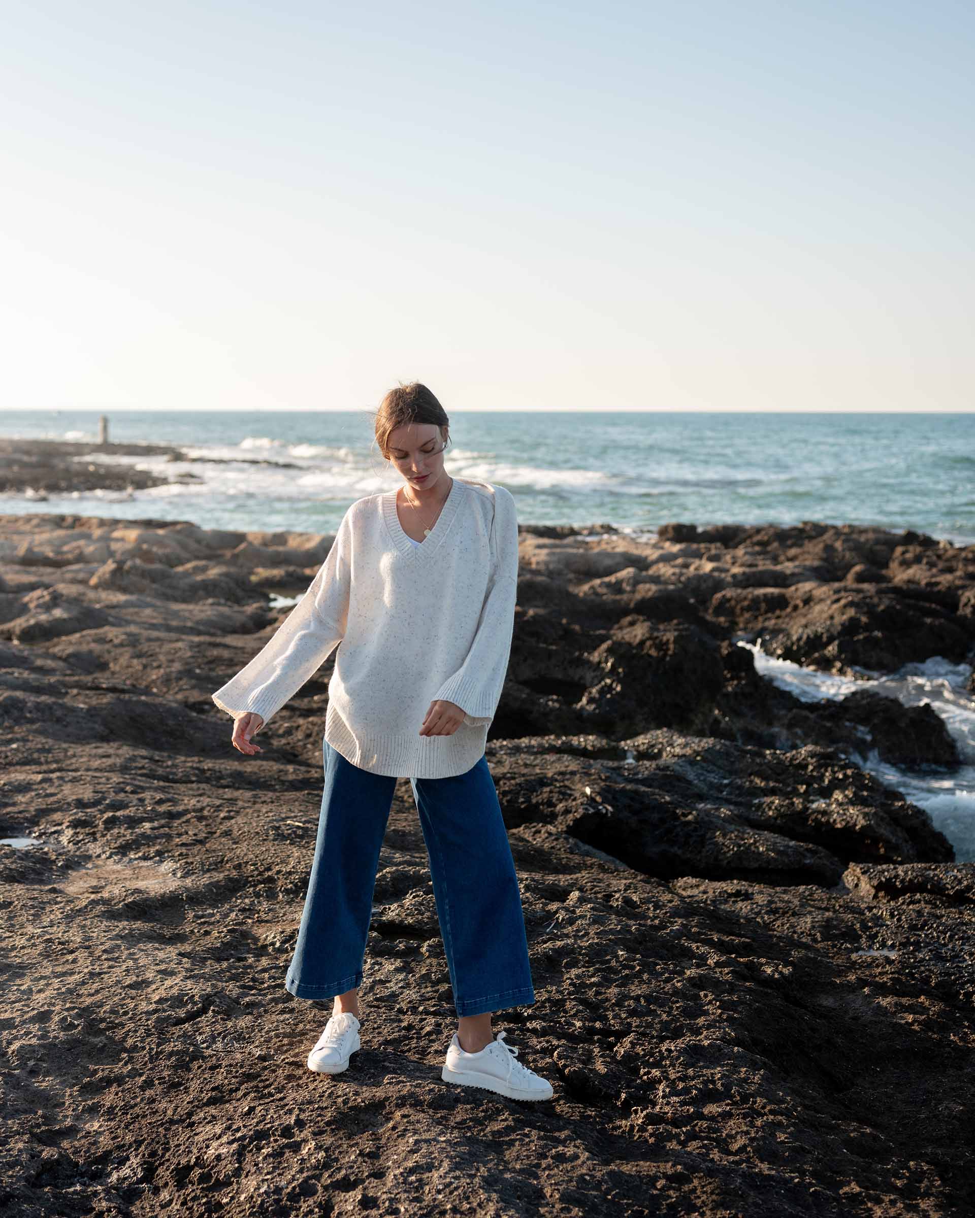 woman wearing mersea montauk v-neck sweater with wide sleeves in cream color standing on rocky beach near water