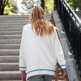 Person wearing a white cardigan with blue trim walking on a sidewalk.