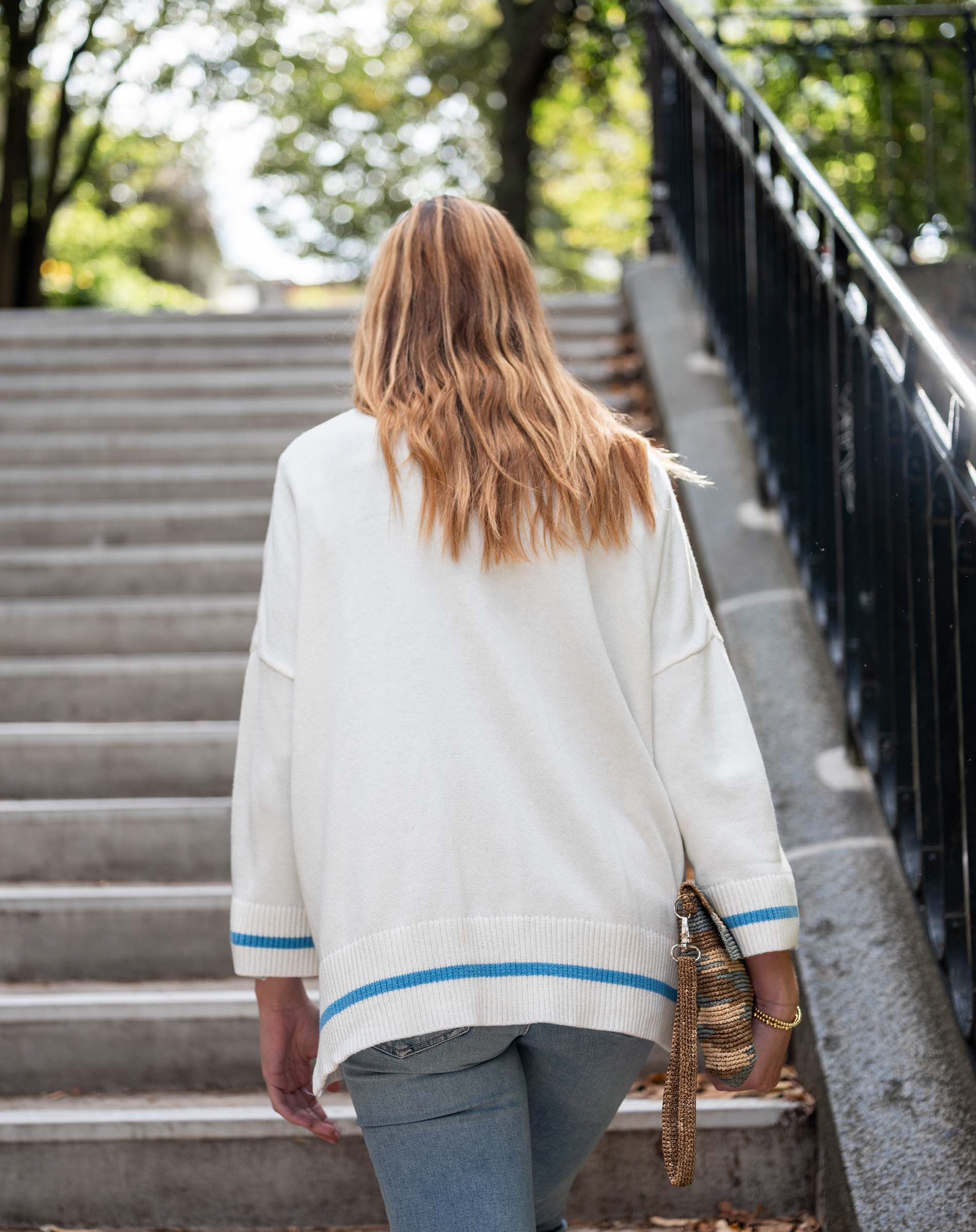 Person wearing a white cardigan with blue trim walking on a sidewalk.