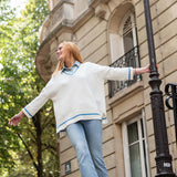 Woman in a white sweater with blue trim standing against a building with trees in the background