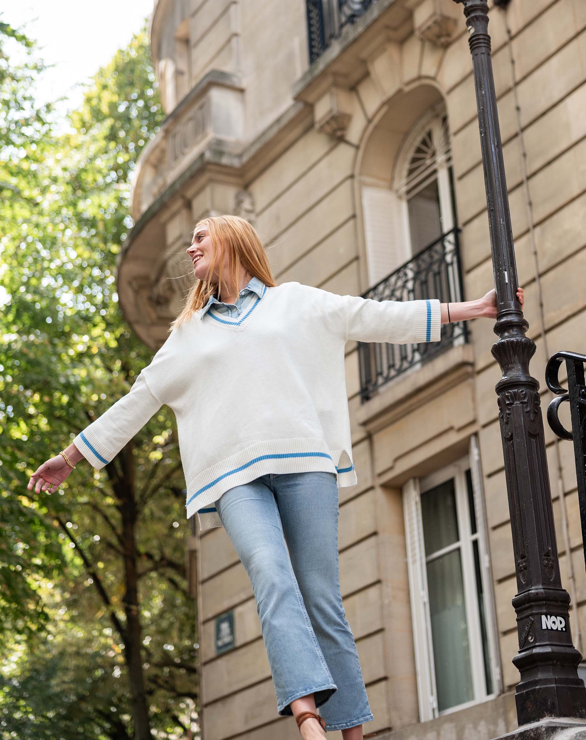 Woman in a white sweater with blue trim standing against a building with trees in the background