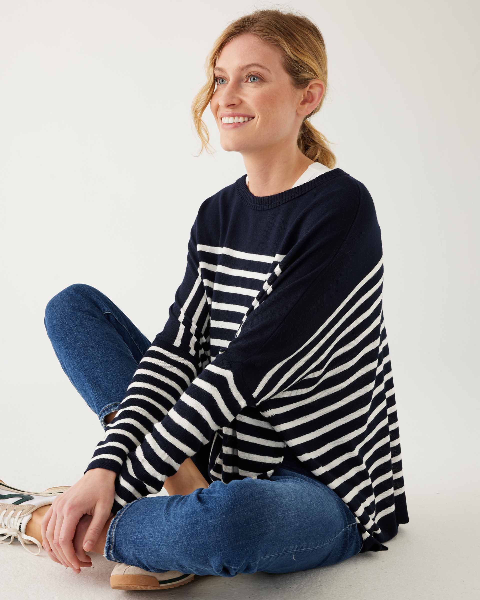 female wearing navy and white striped sweater with blue jeans sitting on a white background